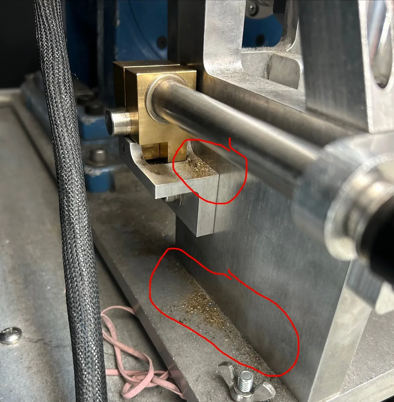 Brass clamp block on the dyno fixture, with two accumulations of brass filings circled in red — one in the tray directly beneath the clamp and one along the base plate below it