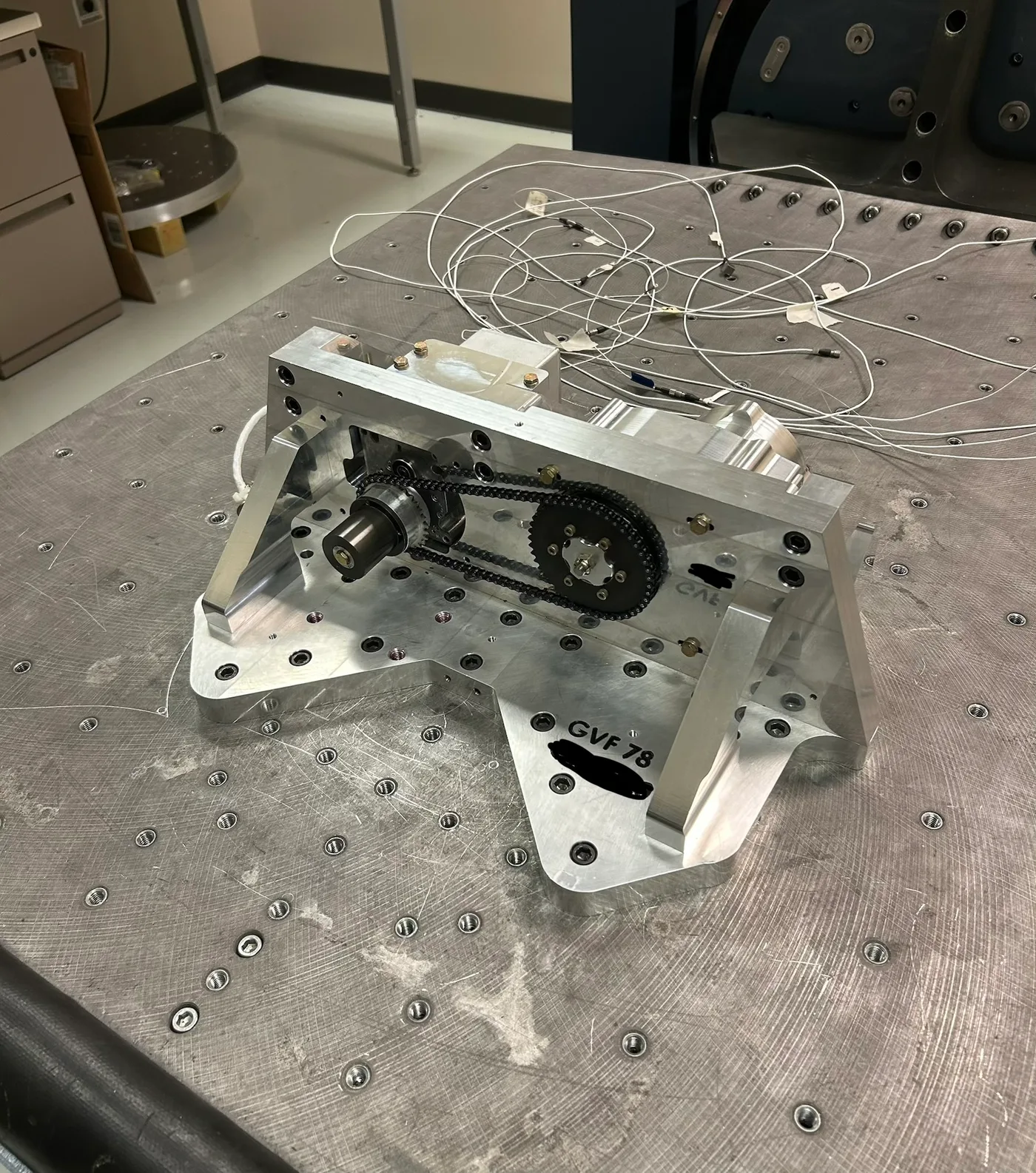 Machined aluminum vibration test fixture bolted to a slip table, with a chain-and-sprocket drive between mounted servos and accelerometer wiring routed off the plate